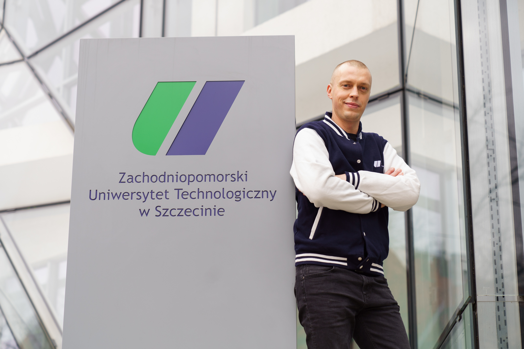 Mężczyzna w granatowo-białej kurtce sportowej stoi z założonymi rękami obok tablicy z logo i nazwą Zachodniopomorskiego Uniwersytetu Technologicznego w Szczecinie, na tle nowoczesnego przeszklonego budynku.