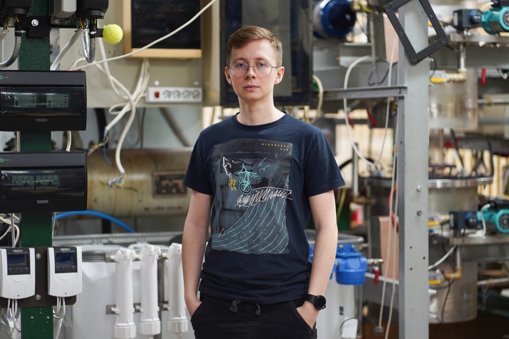 Młody mężczyzna w okularach, ubrany w granatowy T-shirt z kolorową grafiką i czarne spodnie, stoi z rękami w kieszeniach na tle przemysłowego laboratorium pełnego rur, kabli i urządzeń technicznych.